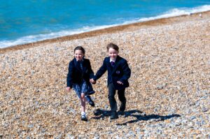 Two children in school uniforms running hand in hand along a pebble beach with the blue sea behind them.